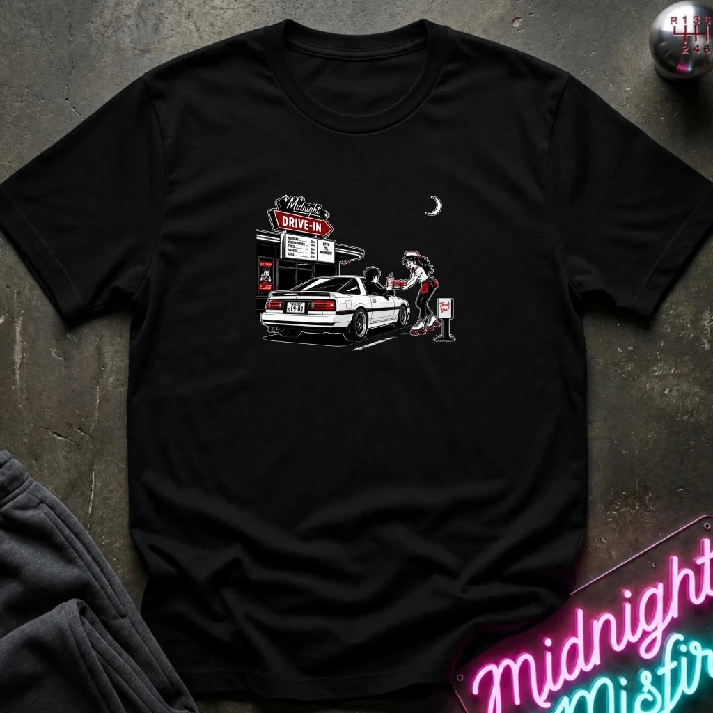 A black short-sleeve t-shirt laid flat on a dark textured surface, featuring a graphic print of a woman pumping gas at a drive-in station next to a white car. 