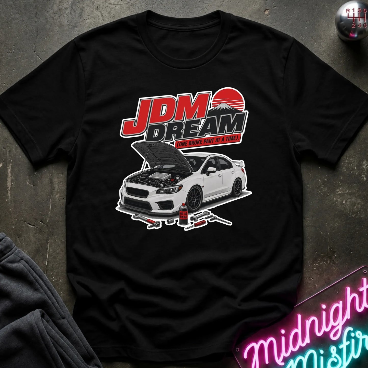 A black short-sleeve graphic T-shirt laid flat on a dark textured surface. The shirt features a large front print with the words 'JDM DREAM' in red, white, and black, above a white sports car with its hood open and a red sun graphic.