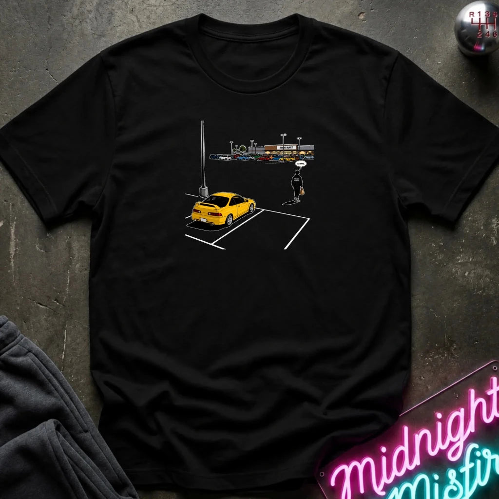 A black short-sleeve t-shirt laid flat on a dark textured surface, featuring a small graphic print of a yellow car, a parking lot, and a lone figure. 
