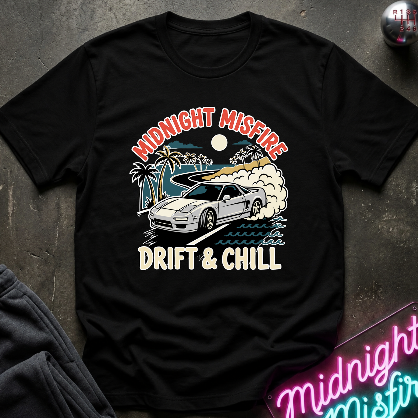 A black short-sleeve graphic T-shirt laid flat on a dark textured surface. The shirt features a colorful retro-style print of a white sports car drifting on water, with palm trees, smoke, and the text 'MIDNIGHT MISFIRE' above and 'DRIFT & CHILL' below.