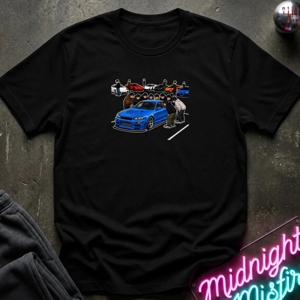 A black short-sleeve graphic t-shirt laid flat on a dark textured surface. The graphic shows a blue sports car with several people and cars in the background.