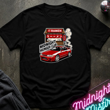 A black short-sleeve graphic t-shirt laid flat on a dark textured surface. The graphic features a red car and a ramen shop with the text 'RAMEN' and 'MIDNIGHT HUNGER CLUB'.