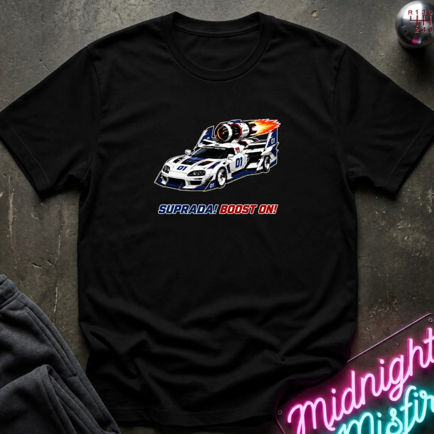 A black short-sleeve t-shirt with a retro-style graphic of a race machine with big boost, displayed flat on a dark textured surface with a neon sign and sweatpants partially visible.