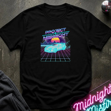 A black short-sleeve t-shirt with a retro 80s synthwave graphic reading 'PROJECT MIDNIGHT' above a neon car and grid scene, laid flat on a dark textured surface with a neon sign and dark pants partially visible.