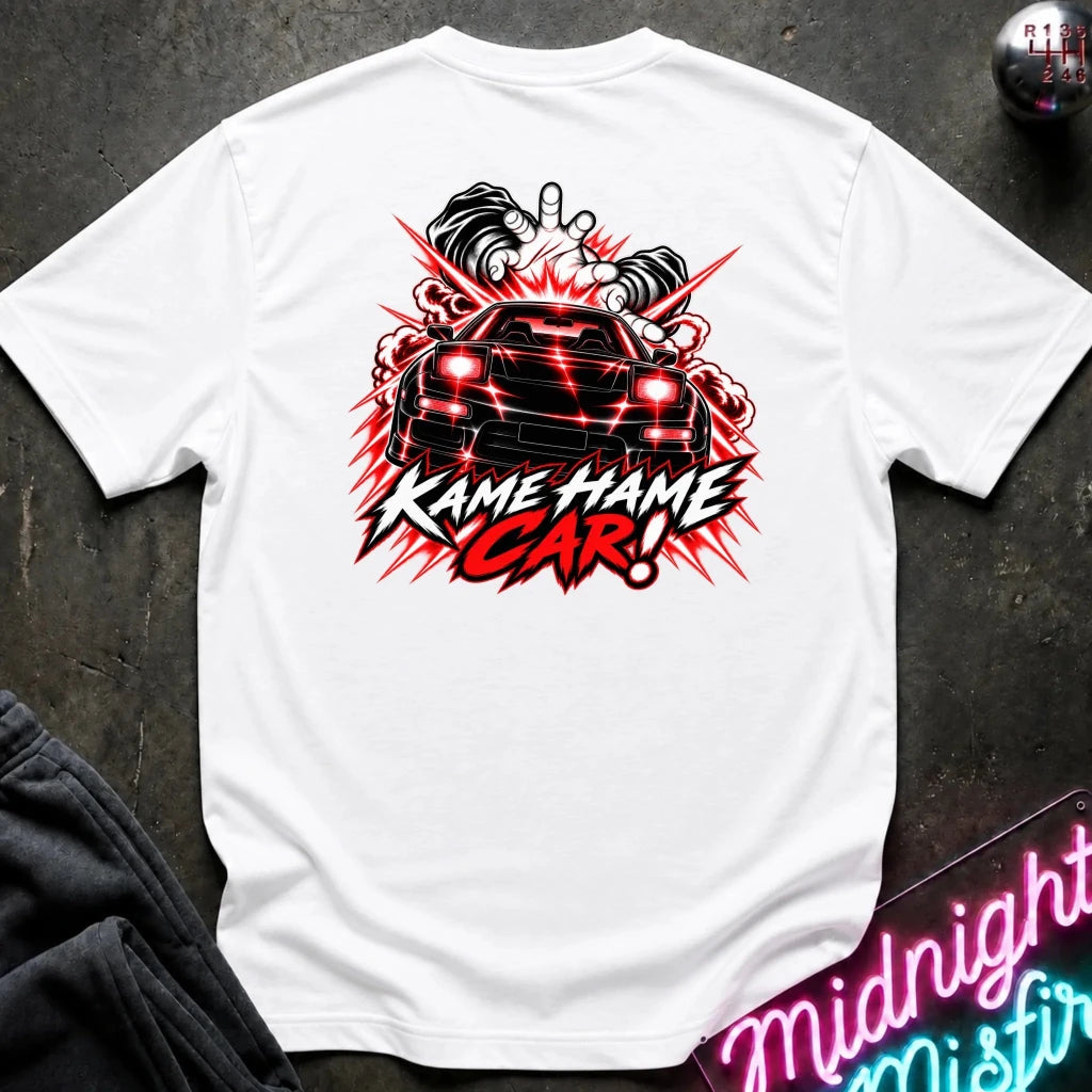 A white short-sleeve t-shirt laid flat on a dark textured surface, featuring a large graphic print of a black sports car with red taillights and the text 'KAME HAME CAR!' 