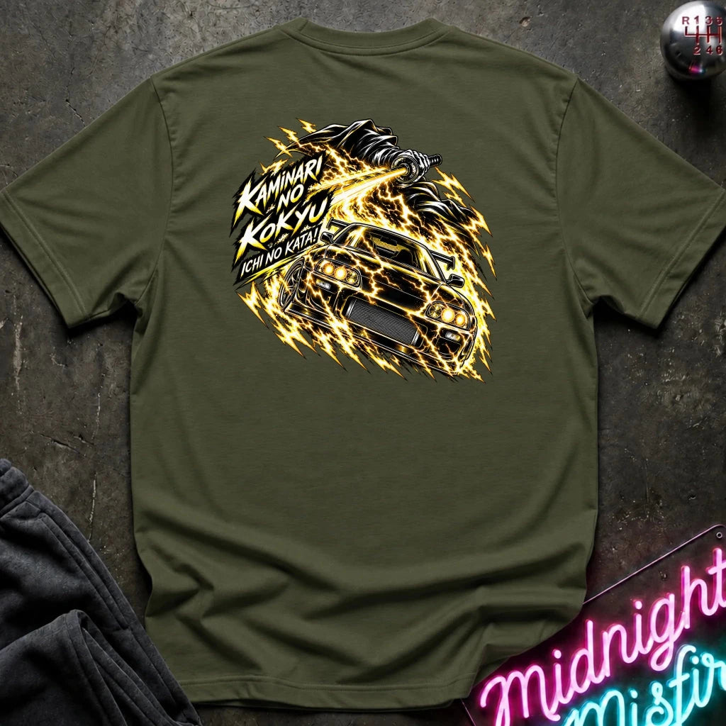 An olive green short-sleeve t-shirt laid flat on a dark textured surface, showing the back graphic with a car and the text "ichi no kata!"