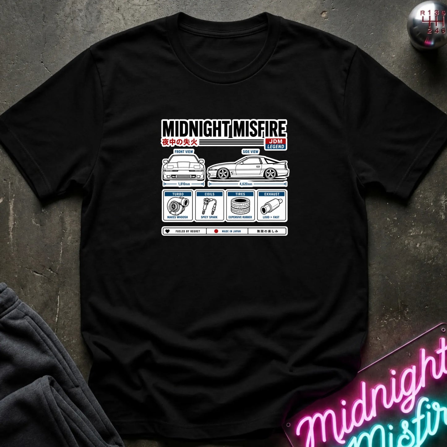 A black short-sleeve graphic t-shirt laid flat on a dark textured surface. The shirt features a white and red graphic print titled 'MIDNIGHT MISFIRE' with a car illustration and small technical-style diagrams.