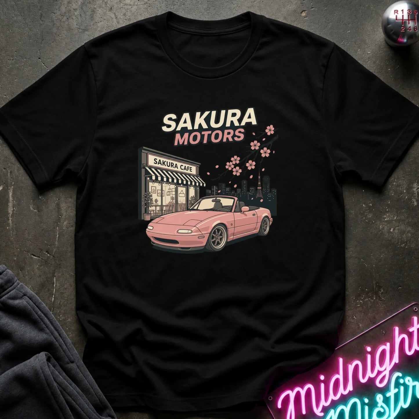 A black short-sleeve graphic T-shirt laid flat on a dark textured surface. The shirt features a retro-style print of a pink convertible car in front of a 'Sakura Motors' cafe, with cream and pink text and cherry blossoms.