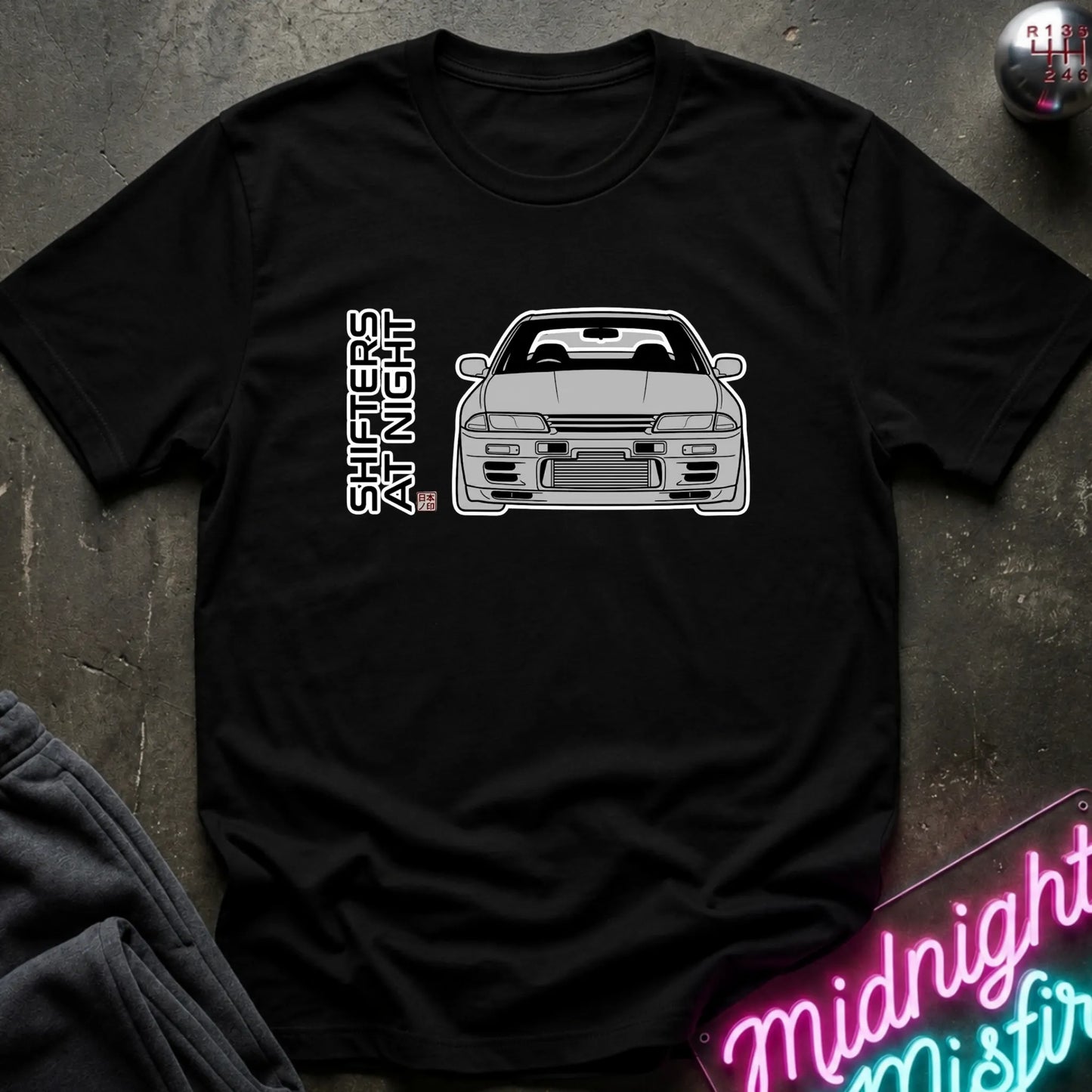 A black short-sleeve t-shirt laid flat on a dark textured surface, featuring a black line art graphic of a sports car and the vertical text 'SHIFTERS AT NIGHT' with a small red and blue flag detail. A neon sign and dark clothing are partially visible as props.