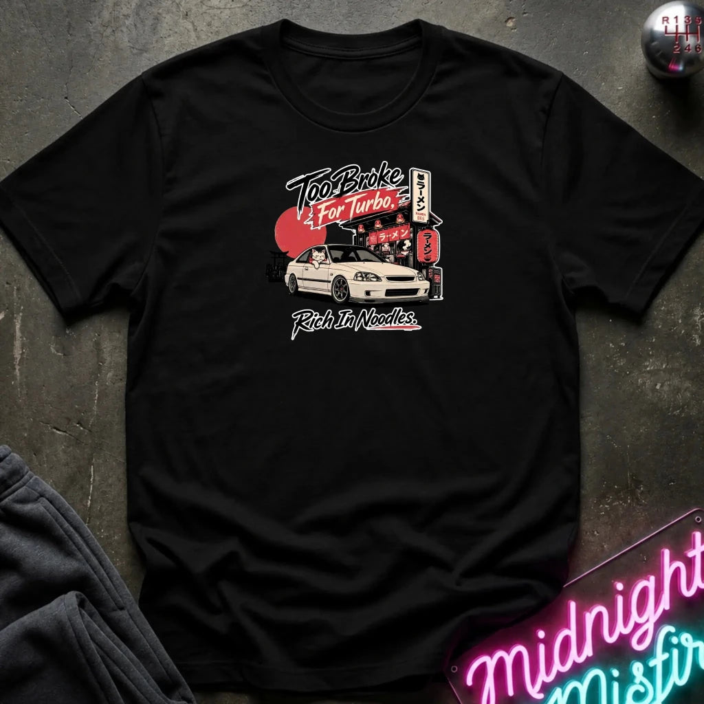 A black short-sleeve graphic t-shirt laid flat on a dark textured surface. The graphic features a white sports car, red and white text reading 'Too Broke For Turbo' and 'Rich In Noodles' with Japanese-style signs.