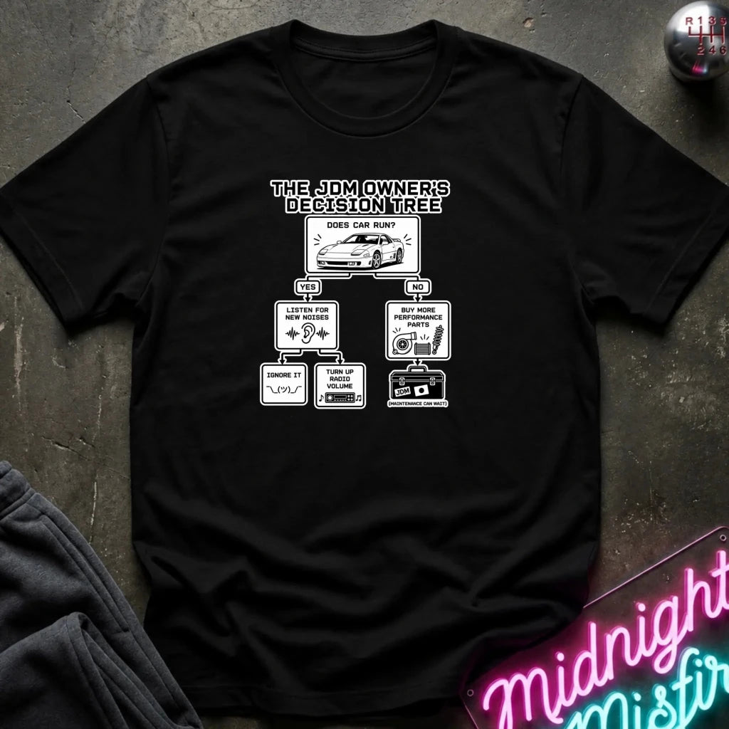 A black short-sleeve graphic T-shirt laid flat on a dark textured surface. The shirt features a white printed decision tree graphic titled 'THE JDM OWNER'S DECISION TREE' with illustrations and text related to car parts and maintenance.