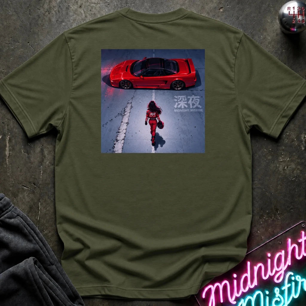 An olive green crewneck t-shirt laid flat on a dark textured surface, featuring a large square graphic print on the back with a red sports car and a figure walking in the snow. A neon sign and folded dark fabric are partially visible in the corners.