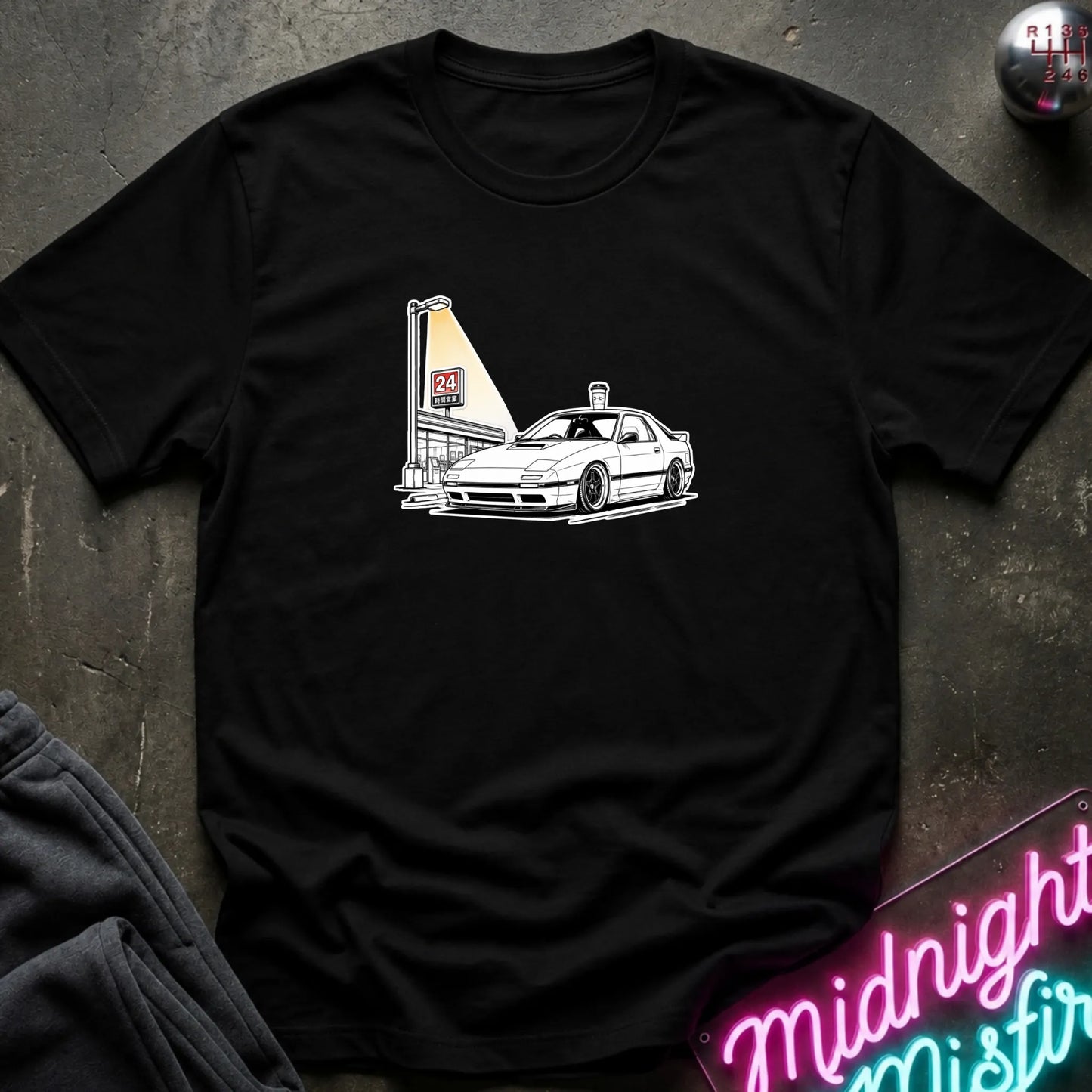 A black short-sleeve crewneck t-shirt laid flat on a dark textured surface, featuring a black line art graphic of a white sports car parked next to a lit street lamp.