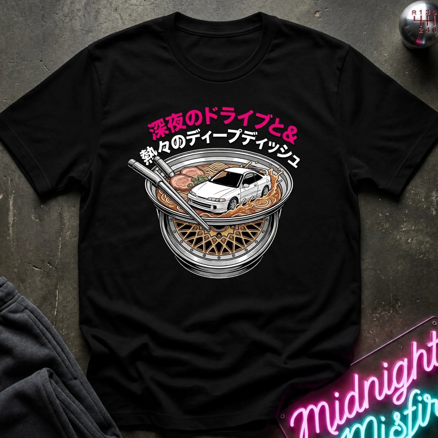 A black short-sleeve graphic T-shirt laid flat on a dark textured surface. The shirt features a large front print with pink and white Japanese text, a white car, and a bowl of ramen with chopsticks.