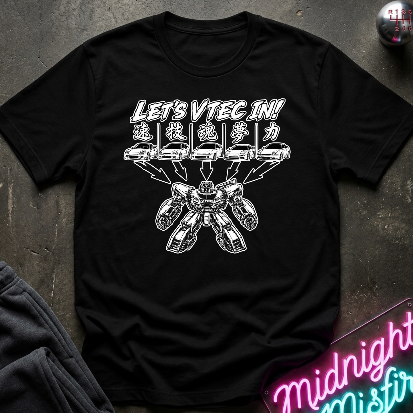 A black short-sleeve graphic t-shirt laid flat on a dark textured surface. The shirt features a white graphic print with the text 'LET'S VTEC IN!' and stylized car illustrations.