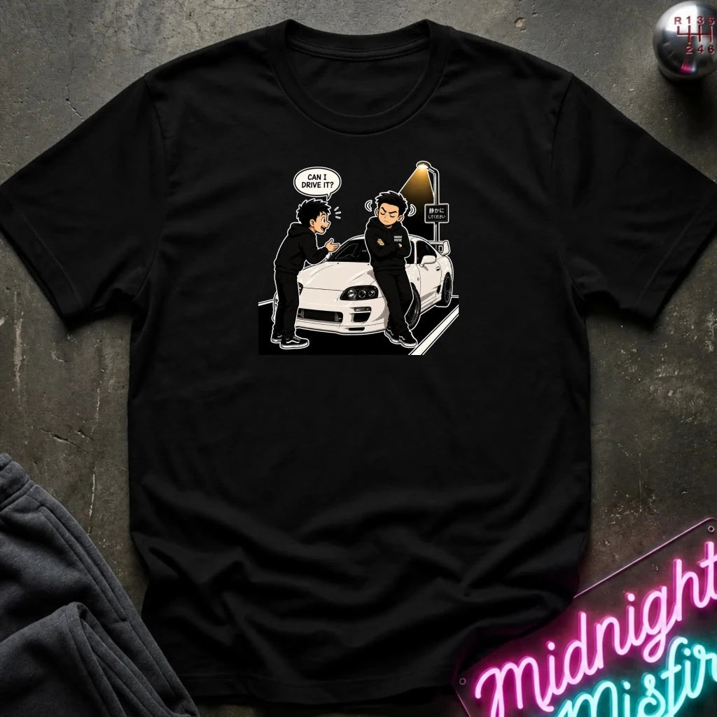 A black short-sleeve graphic t-shirt laid flat on a dark textured surface. The graphic shows a white sports car with two cartoon figures standing beside it, one with a speech bubble 