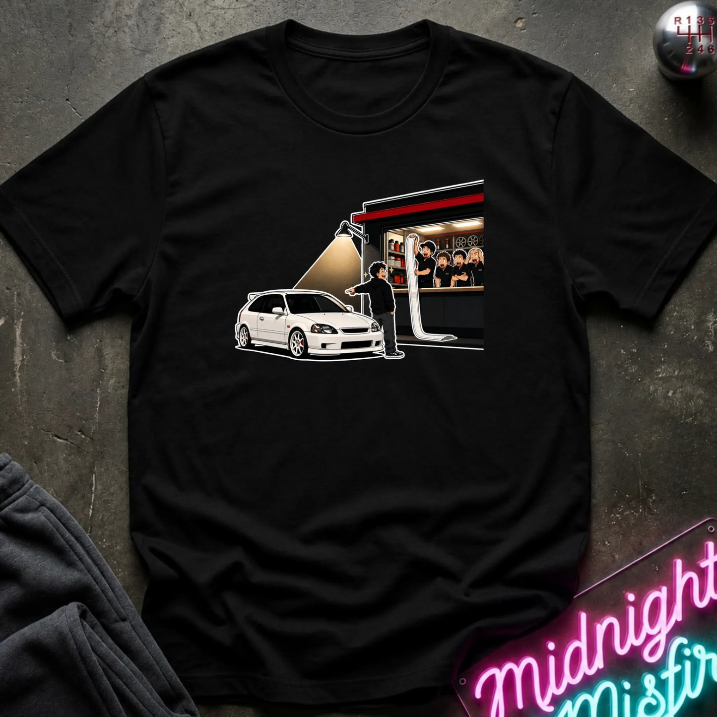 A black short-sleeve graphic t-shirt laid flat on a dark textured surface. The graphic shows a white hatchback car next to a shop with a spotlight effect.