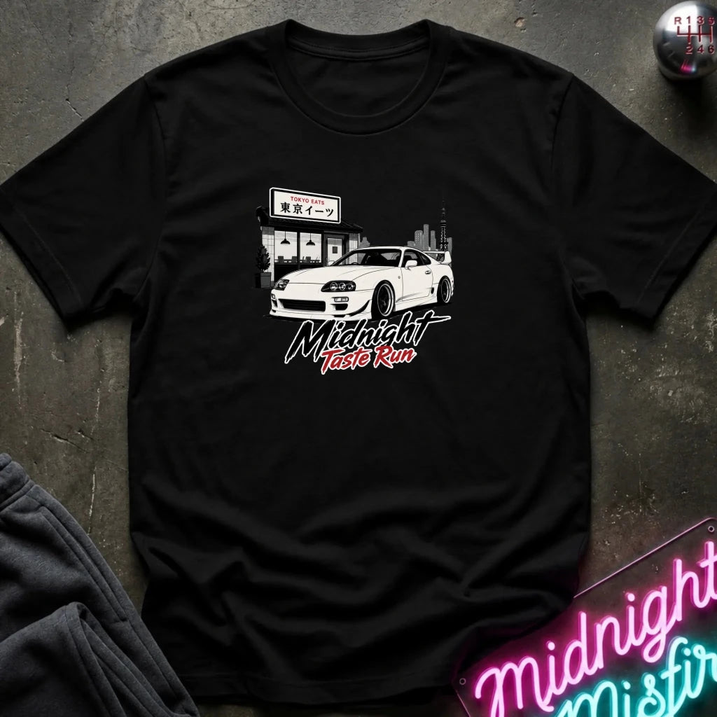 A black short-sleeve graphic t-shirt laid flat on a dark textured surface. The shirt features a white sports car graphic with Japanese text and the words 'Midnight Taste Run' in stylized white and red lettering.