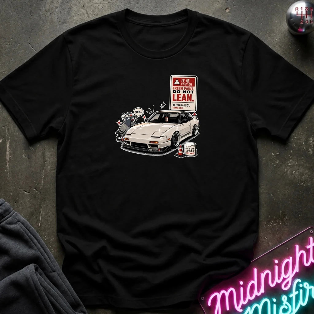 A black short-sleeve graphic t-shirt laid flat on a dark textured surface. The graphic features a white sports car with a 'DO NOT LEAN' sign and a cartoon character.
