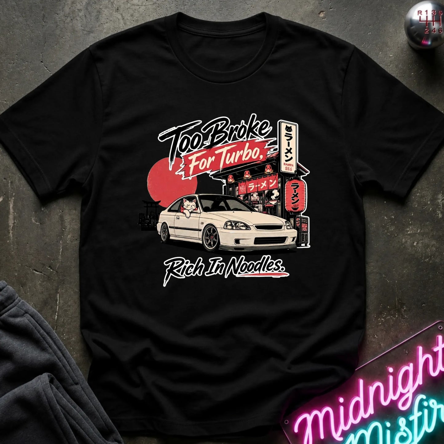 Black short-sleeve graphic T-shirt laid flat on a dark textured surface. The shirt features a printed design of a white sports car in front of a Japanese-style street scene with a red sun and neon signs, plus the text 'Too Broke For Turbo' and 'Rich In Noodles.
