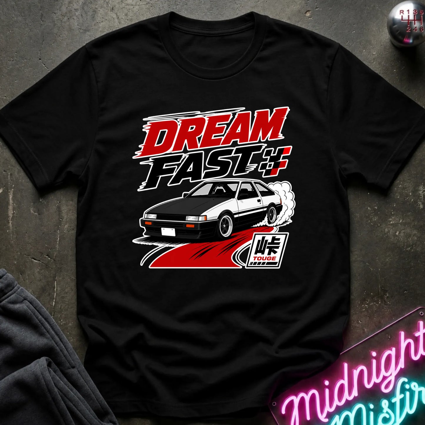 A black short-sleeve graphic T-shirt laid flat on a dark textured surface. The shirt features a large front print with the words “DREAM FAST” in red, white, and black, above an illustration of a black-and-white sports car with smoke and a stylized logo.