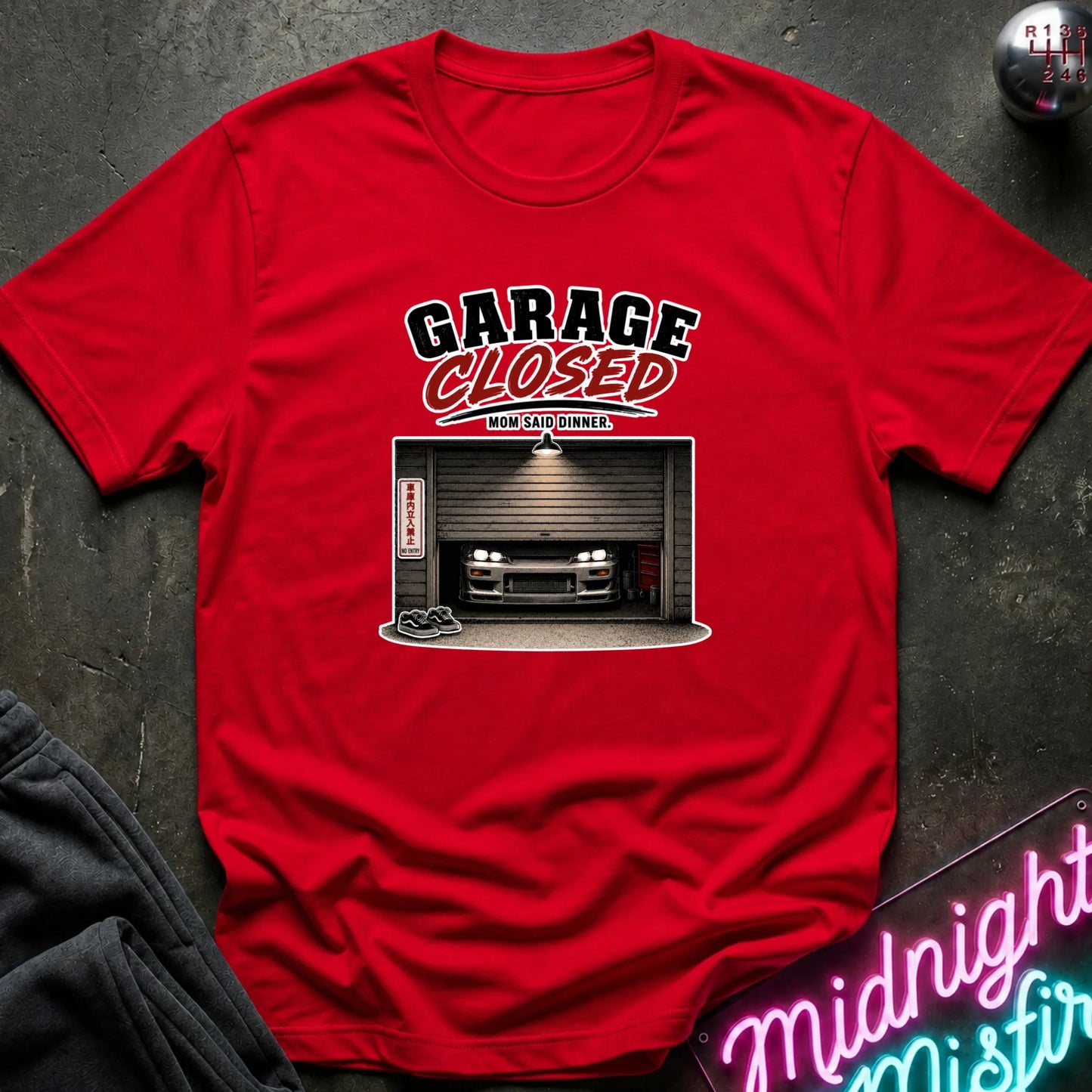 A red short-sleeve graphic t-shirt laid flat on a dark textured surface. The shirt features a graphic print with the text 'GARAGE CLOSED' and an illustration of a car inside a garage.