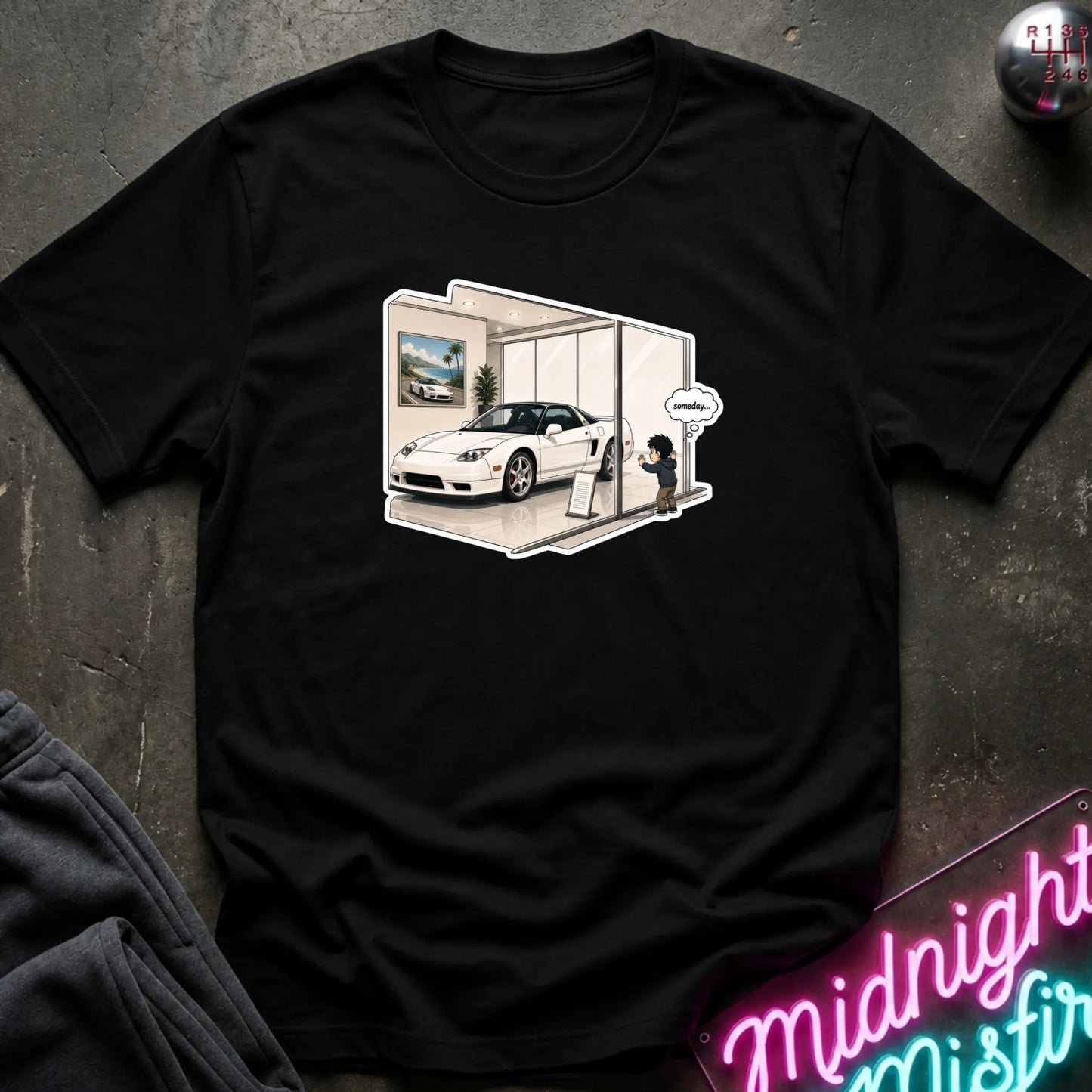 A black short-sleeve graphic t-shirt laid flat on a dark textured surface. The graphic shows a white sports car inside a showroom with a small cartoon character and sign.