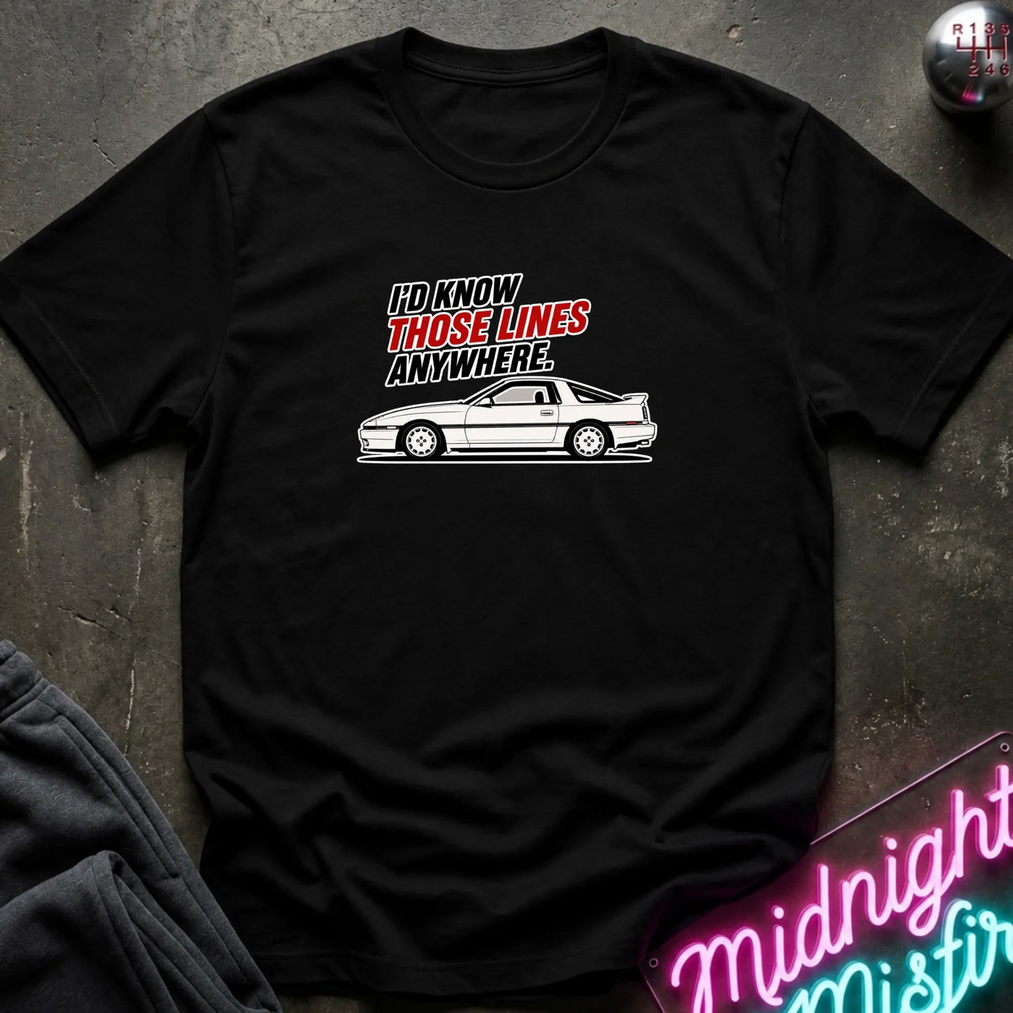 A black short-sleeve graphic t-shirt laid flat on a dark textured surface. The shirt features a white line art drawing of a sports car and the text 'I'D KNOW THOSE LINES ANYWHERE.