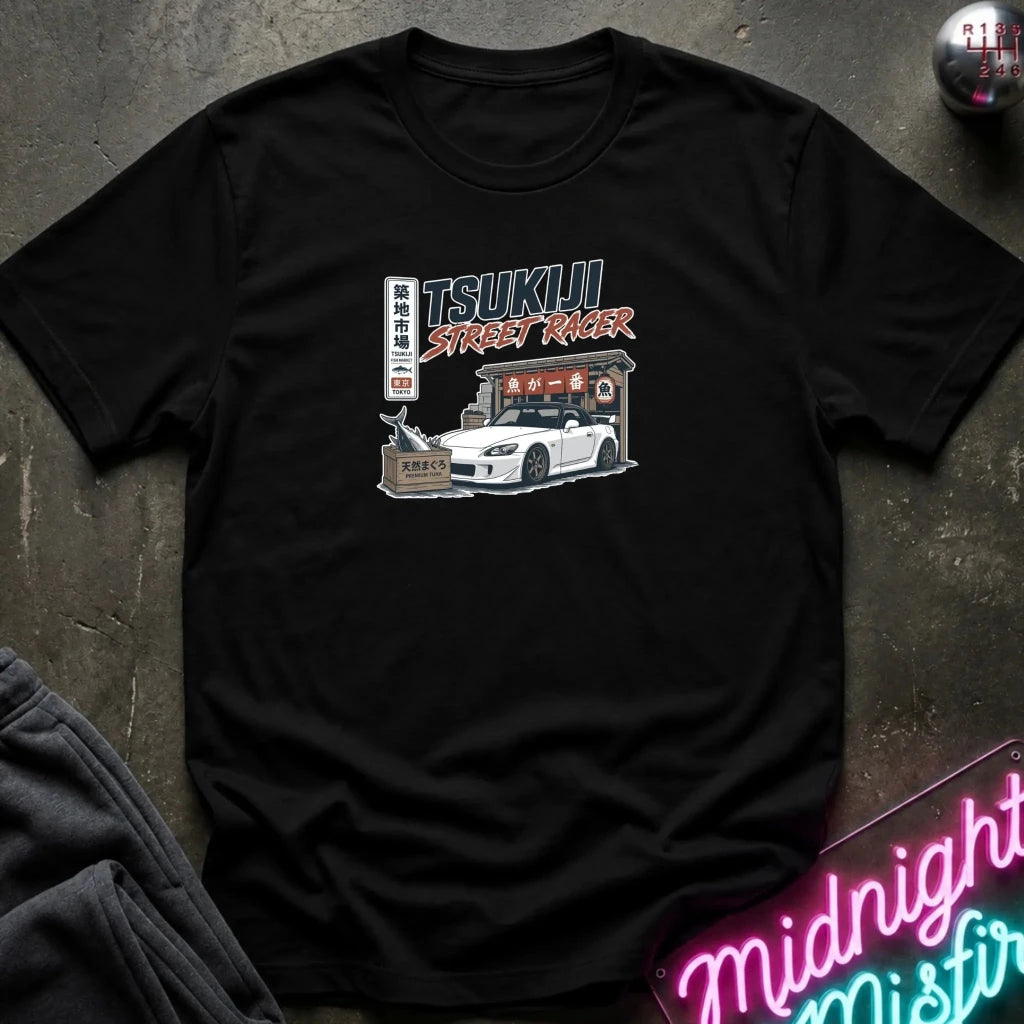 A black short-sleeve graphic t-shirt laid flat on a dark textured surface. The graphic features a white sports car, Japanese text, and a building illustration with 'TSUKIJI STREET RACER' written above it.