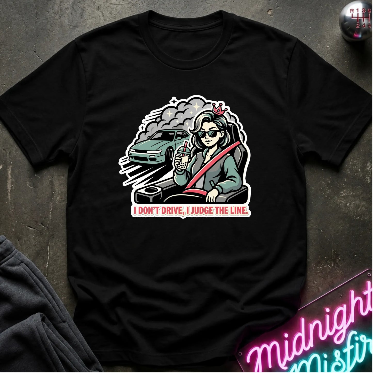 A black short-sleeve graphic t-shirt laid flat on a dark textured surface. The graphic shows a cartoon woman wearing sunglasses and a seatbelt with a teal car behind her, above the text 'I DON'T DRIVE, I JUDGE THE LINE.