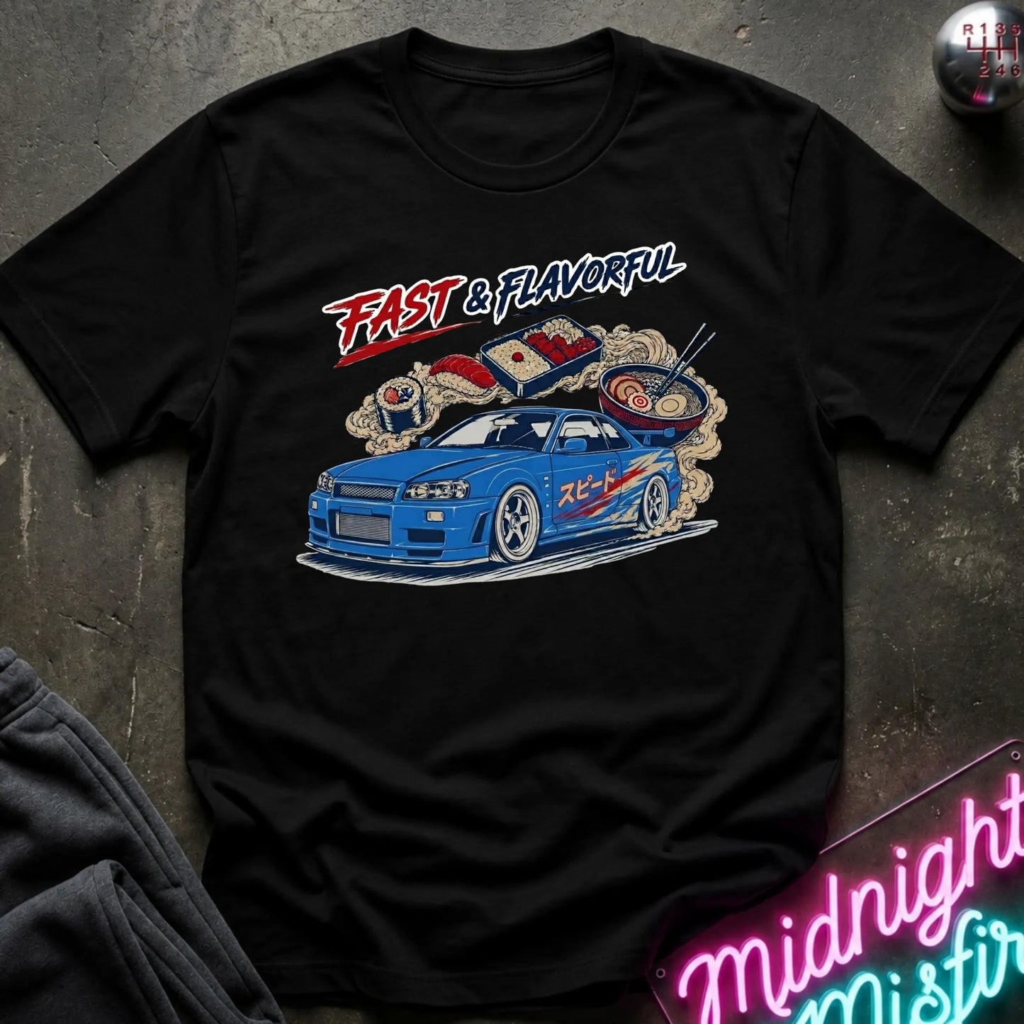 A black short-sleeve graphic T-shirt laid flat on a dark textured surface. The shirt features a colorful front print with the words “FAST & FLAVORFUL” above a blue race car surrounded by ramen noodles and steam.