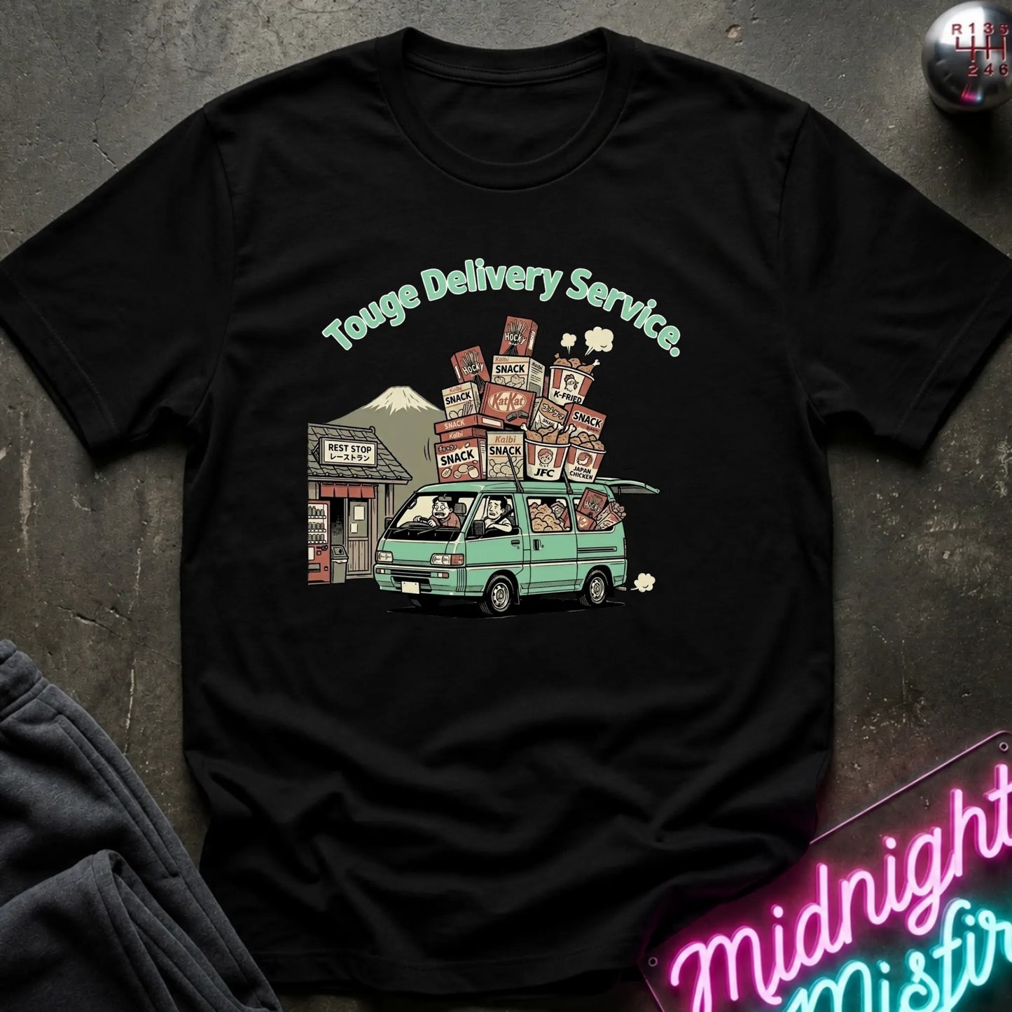 A black short-sleeve graphic t-shirt laid flat on a dark textured surface. The shirt features a colorful cartoon-style print of a mint green van carrying stacked boxes, with the text 'Touge Delivery Service.