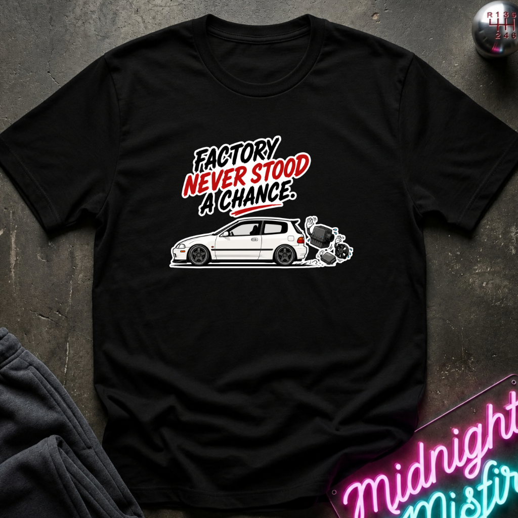 A black short-sleeve graphic t-shirt laid flat on a dark textured surface. The shirt features a white car illustration and the text 'FACTORY NEVER STOOD A CHANCE' in red and white lettering.