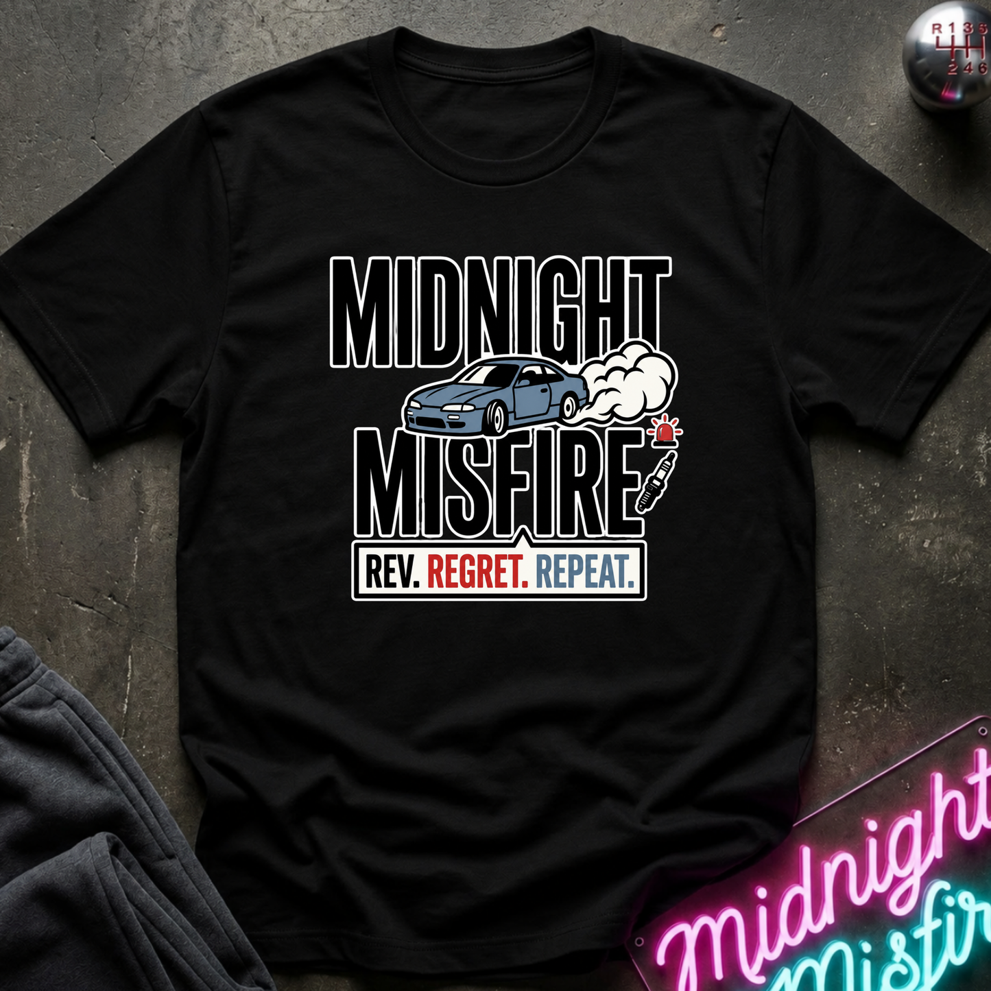 A black short-sleeve graphic T-shirt laid flat on a dark textured surface. The shirt features a distressed white and blue print reading 'MIDNIGHT MISFIRE' with a blue car, smoke, and a small icon.