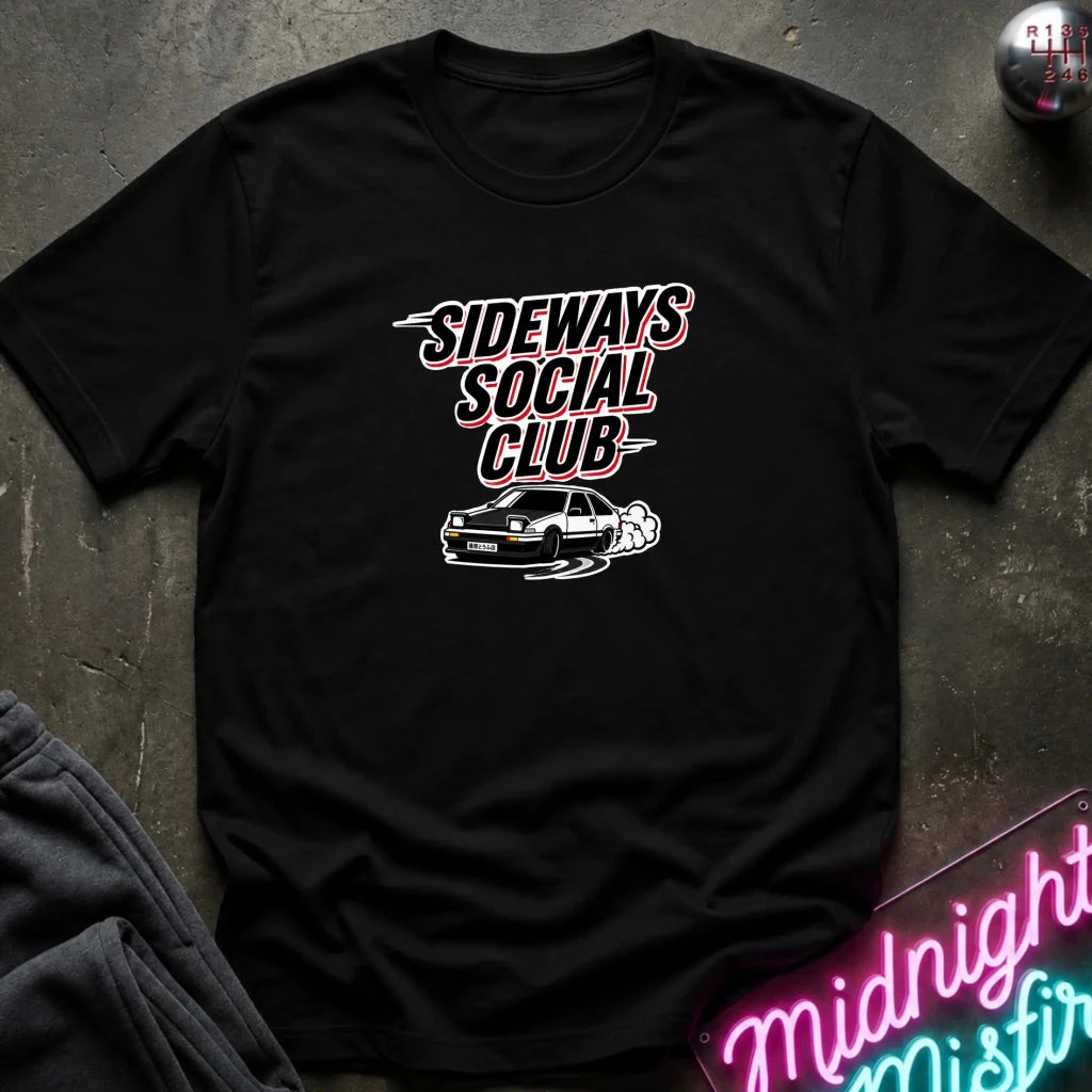 A black short-sleeve graphic t-shirt laid flat on a dark textured surface. The shirt features a white and pink graphic that reads 'SIDeways Social Club' above a white car illustration.
