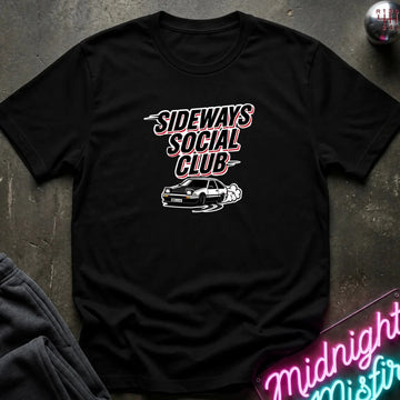 A black short-sleeve graphic t-shirt laid flat on a dark textured surface. The shirt features a white and pink graphic that reads 'SIDeways Social Club' above a white car illustration.