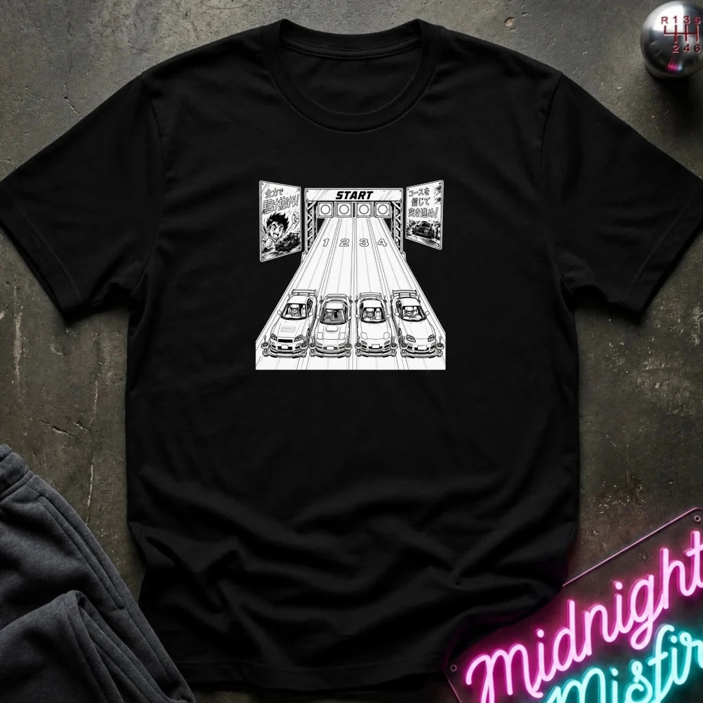 A black crewneck t-shirt featuring a black and white graphic print of 4 cars lined up at a race starting line with the word 'START' above them. The shirt is laid flat on a dark textured surface with a neon sign and dark pants partially visible in the corners.