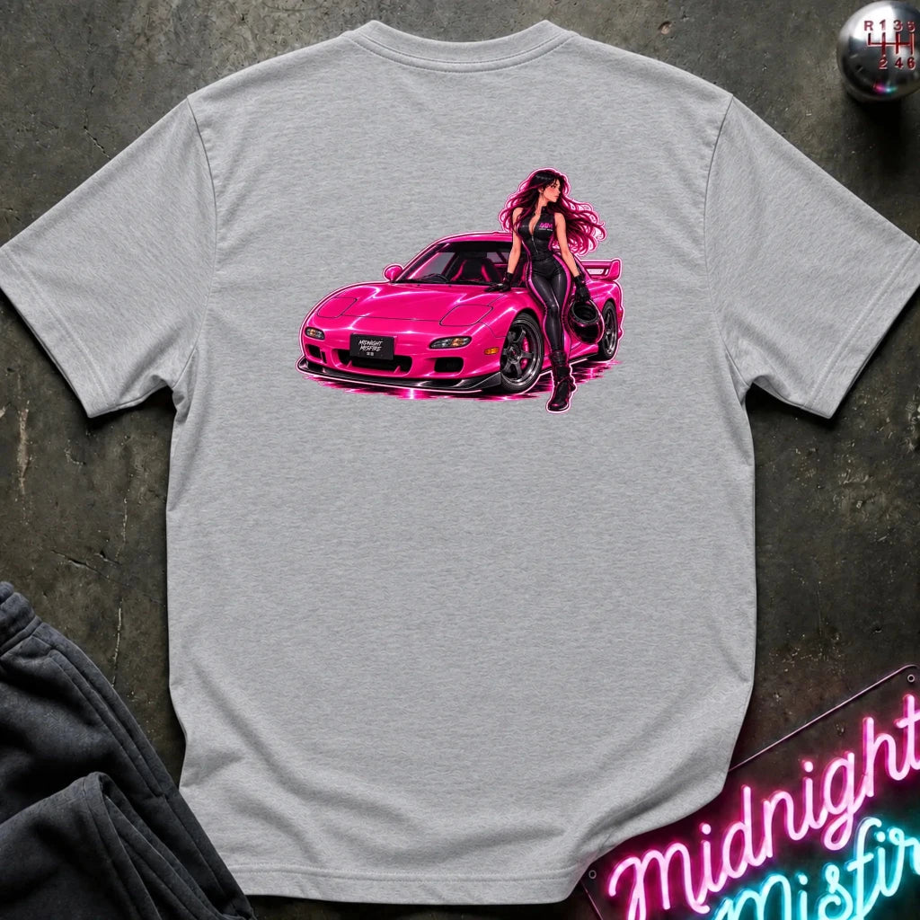 A light gray heather crewneck t-shirt featuring a graphic print of a woman with long dark hair posing next to a bright pink sports car. The shirt is displayed flat on a dark textured surface with a neon sign and sweatpants partially visible.