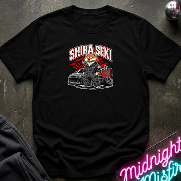 A black short-sleeve graphic T-shirt laid flat on a dark textured surface. The shirt features a central graphic of a fox character in a suit standing next to a car and tool chest, with the text 'SHIBA SEKI' above.