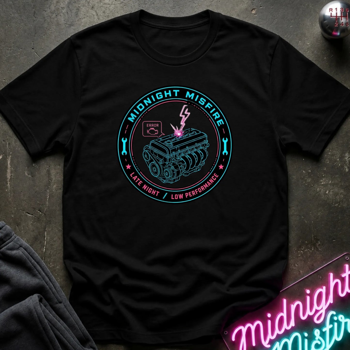 A black short-sleeve graphic T-shirt laid flat on a dark textured surface. The shirt features a circular teal and pink graphic with an engine illustration and the words 'MIDNIGHT MISFIRE' and 'LATE NIGHT / LOW PERFORMANCE.