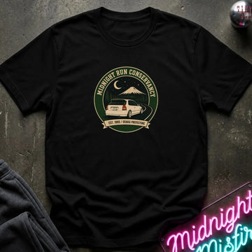 A black short-sleeve t-shirt laid flat on a dark textured surface, featuring a circular vintage-style graphic on the chest with a white car, mountain, moon, and the words 'Midnight Run Conservancy'