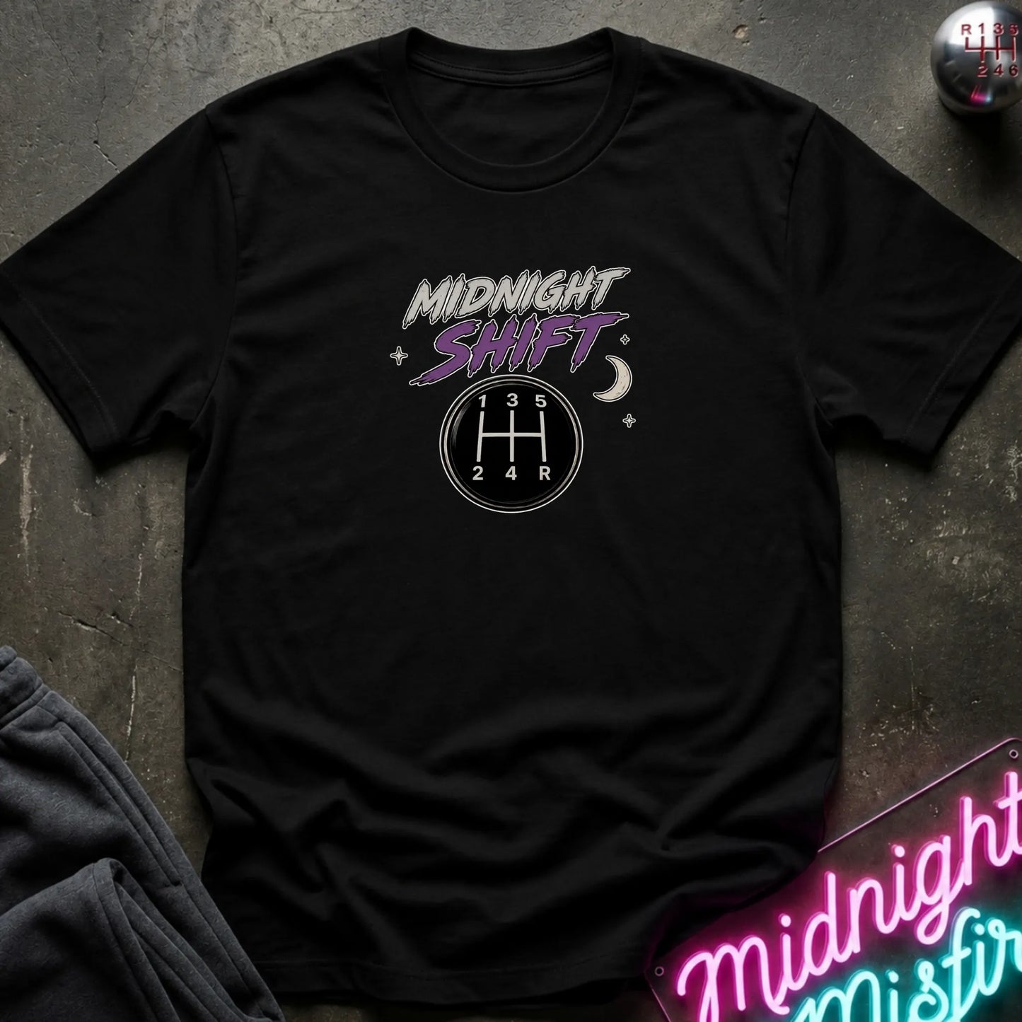 A black short-sleeve graphic t-shirt laid flat on a dark textured surface. The shirt features a 'Midnight Shift' graphic with a 5-speed manual gearshift pattern and stars in white and purple.