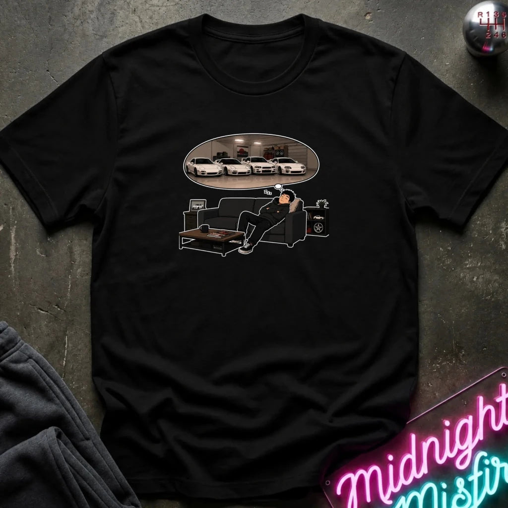A black short-sleeve graphic t-shirt laid flat on a dark textured surface. The graphic shows a man sitting on a black couch dreaming, with an oval reflection of several white cars above him.