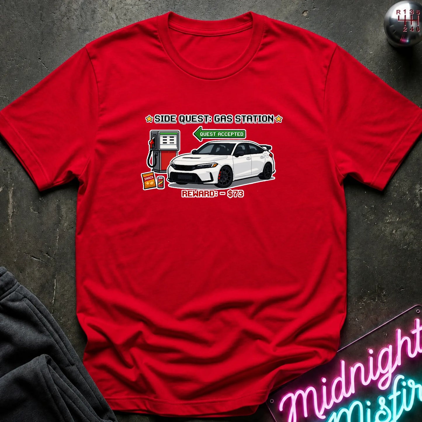 A red short-sleeve graphic T-shirt laid flat on a dark textured surface. The shirt features a printed design of a white sports car at a gas station with text reading 'SIDE QUEST GAS STATION'.