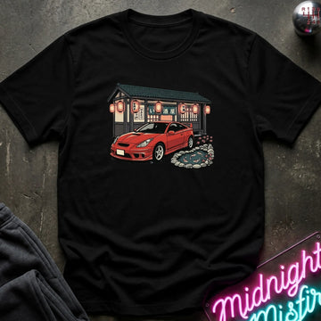 A black short-sleeve crewneck t-shirt laid flat on a dark textured surface, featuring a graphic print of a red sports car parked in front of a Japanese-style building with red lanterns.