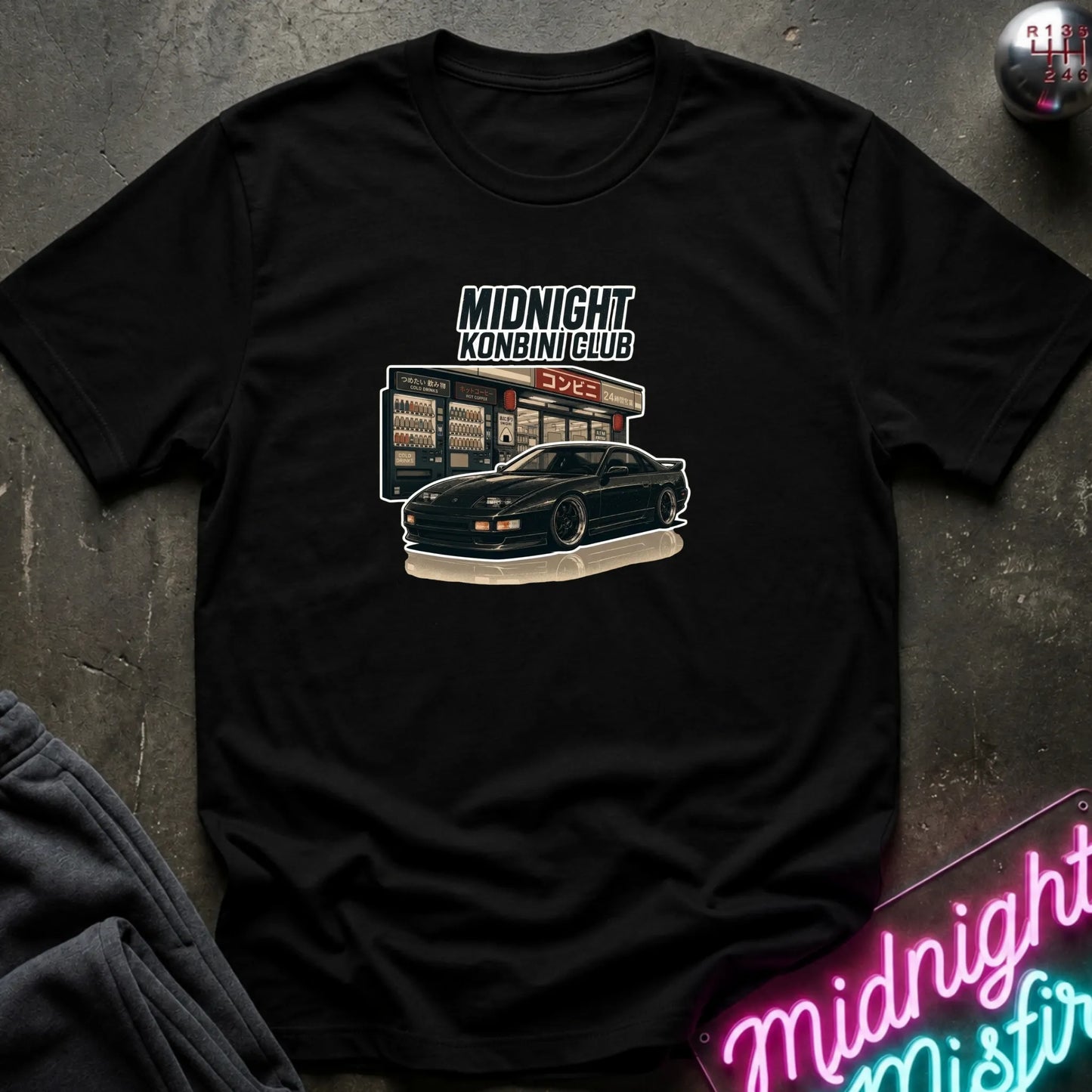 A black short-sleeve graphic t-shirt laid flat on a dark textured surface. The graphic features the text 'MIDNIGHT KONBINI CLUB' above an illustration of a black sports car parked in front of a convenience store.