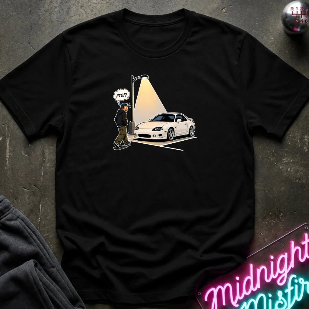A black short-sleeve graphic t-shirt laid flat on a dark textured surface. The graphic shows a white sports car parked under a spotlight with a small figure and a speech bubble 