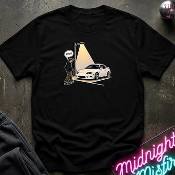 A black short-sleeve graphic t-shirt laid flat on a dark textured surface. The graphic shows a white sports car parked under a spotlight with a small figure and a speech bubble 