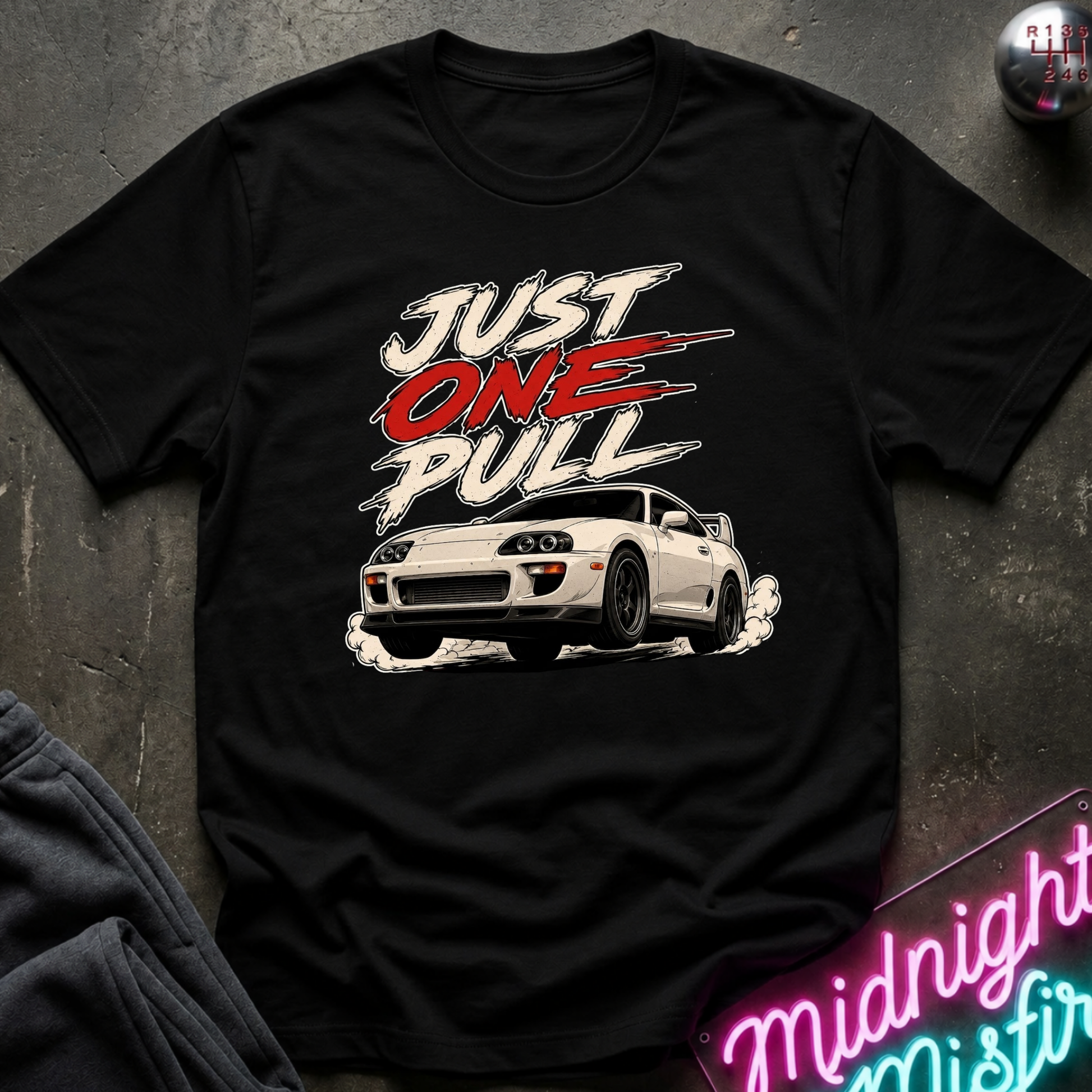A black short-sleeve graphic t-shirt laid flat on a dark textured surface. The shirt features a white sports car and the text 'JUST ONE PULL' in white and red.