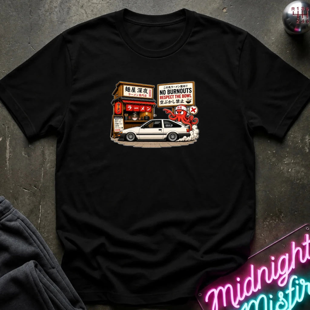 A black short-sleeve t-shirt laid flat on a dark textured surface, featuring a graphic print of a white sports car parked in front of a Japanese street shop with signs reading 'NO BURNOUTS'.